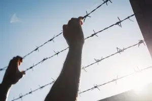 Hands reaching for barbed wire fence against sky.